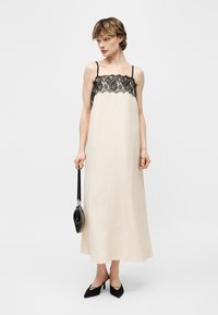 Femeie purtând o rochie-slip crem cu dantelă neagră, ținând o poșetă mică neagră și purtând pantofi cu toc ascuțit negri, pe un fundal simplu.