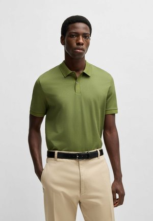 Olivgrünes Polohemd mit kurzen Ärmeln, ausgestattet mit einem strukturierten Stoff und einer Knopfleiste mit zwei Knöpfen, kombiniert mit beigen Hosen und einem schwarzen Gürtel.