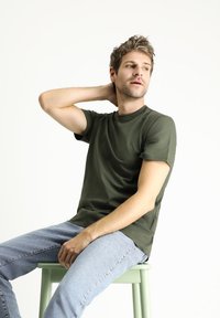 T-shirt verde oliva con maniche corte e scollatura a girocollo, abbinata a jeans azzurri. Il modello è seduto su uno sgabello verde.