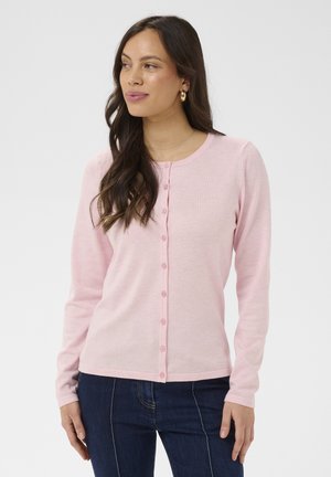 Kobieta o długich, ciemnych włosach, nosząca złote kolczyki koła, jasnoróżowy sweter zapinany na guziki oraz ciemnoniebieskie jeansy, patrząca na bok.