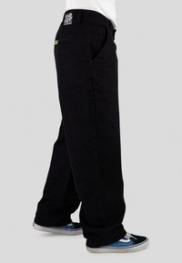 Black denim pants with a relaxed fit, featuring front pockets and a branded waistband tag. Worn with blue and black sneakers.