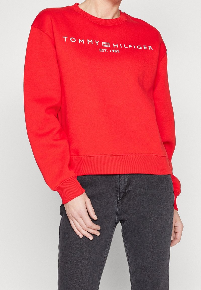 Personne portant un sweat-shirt Tommy Hilfiger rouge vif avec texte blanc du logo et jean gris foncé, mains reposant près des hanches.