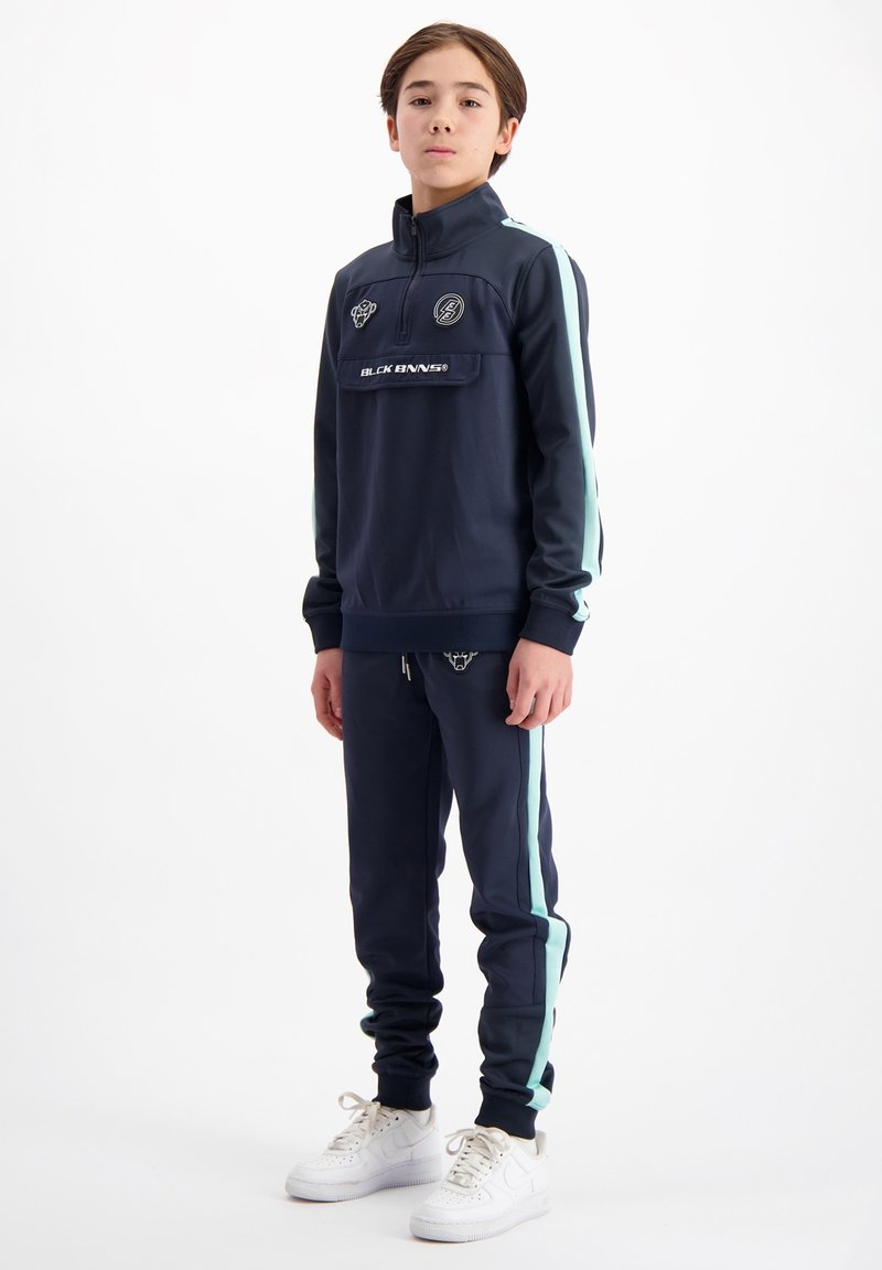 Donkerblauwe trainingsoutfit met lichte blauwe zijstrepen, featuring een ritskraag en een voorzak. Gecombineerd met witte sneakers. Gladde stoftextuur.