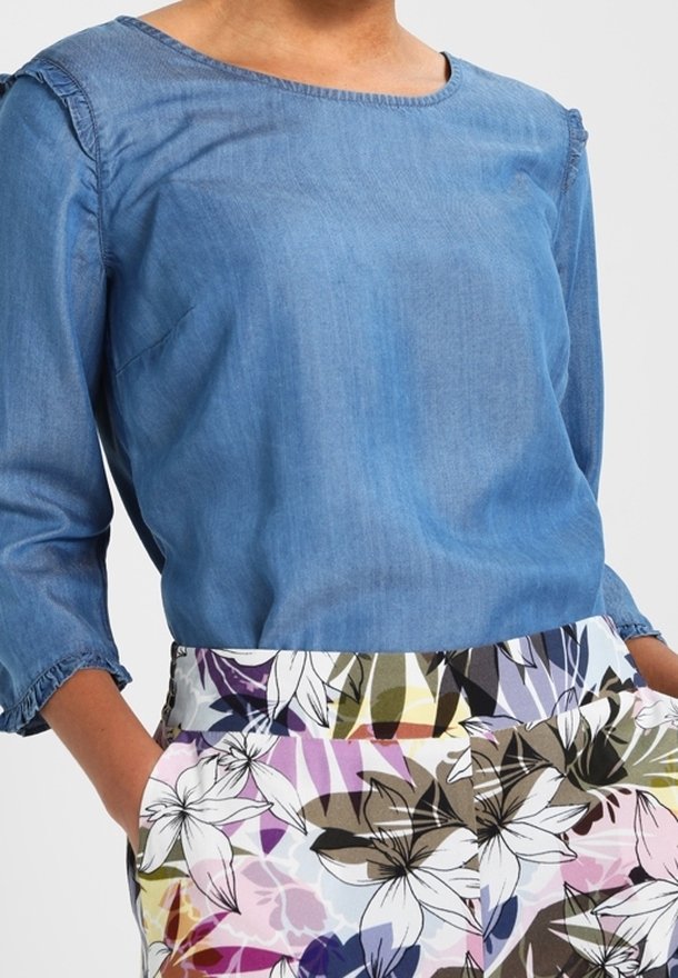 Femme portant un chemisier bleu à manches longues associé à un pantalon taille haute à motifs floraux multicolores avec des lys blancs.