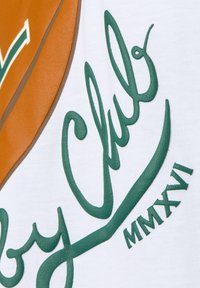 Biała tkanina z teksturowanym zielonym napisem "by Club" oraz "MMXVI", obok gładkiego brązowego elementu dekoracyjnego.