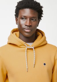 Felpa con cappuccio color giallo senape, con una finitura texturizzata, dotata di un laccio blu e bianco e di un piccolo logo di un albero nero sul petto.