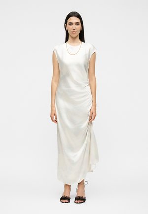 Γυναίκα με ίσια σκούρα μαλλιά που φοράει λευκό σατέν μίντι φόρεμα χωρίς μανίκια, χρυσό κολιέ αλυσίδα και μαύρα δαντελένια πέδιλα με ψηλό τακούνι.