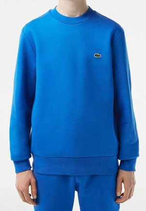 Homme portant un sweat-shirt bleu à manches longues et un pantalon bleu assorti avec un petit logo crocodile vert sur la poitrine.