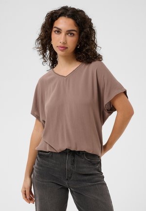 Donna con capelli ricci che indossa una camicetta morbida color malva con scollo a V e jeans neri, in piedi con una mano sui fianchi davanti a uno sfondo uniforme.