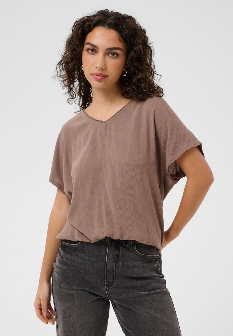 Femme aux cheveux bouclés portant un chemisier ample mauve à col en V et un jean noir, debout avec une main sur la hanche sur un fond uni.