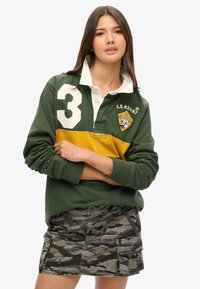 Grünes Rugby-Shirt mit einem gelben Streifen und der weißen Nummer 3. Kombiniert mit einem Rock im Camouflage-Muster, der Taschen und einen Knopfverschluss hat.