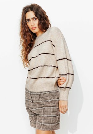Femme aux longs cheveux bouclés portant un pull beige rayé et un short taille haute à carreaux marron, debout devant un fond blanc.