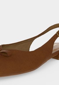 Καφέ δερμάτινο σανδάλι με επίπεδη σόλα, μικρό φιόγκο στη μύτη και χρυσή αγκράφα στο λουράκι του αστραγάλου, σε απλό φόντο.