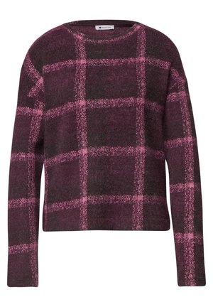 Kvinde set fra siden iført en lilla og lyserød ternet sweater og lyseblå jeans mod en ensfarvet lys baggrund.