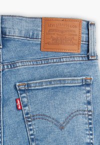 Světle modré džíny z denimu s hnědým koženým sloupkem s vyraženým textem, oranžovým prošíváním a malou červenou nášivkou na zadní straně.