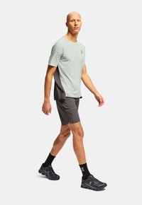 Jasnozielona koszulka sportowa z ciemnoszarymi bokami, połączona z szarymi szortami i czarnymi skarpetkami. Obuwie ma elegancki design z ciemnymi akcentami.