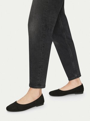 Piernas con jeans ajustados negros y bailarinas de ante negro sobre un fondo claro.