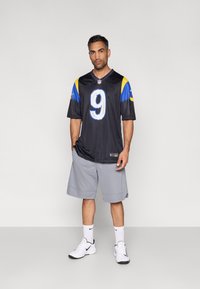Černý fotbalový dres s modrými a žlutými akcenty, číslo 9, doplněný světle šedými šortkami a bílými teniskami.