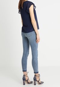Blusa de gasa marina con mangas acampanadas, combinada con jeans skinny de color azul claro. El calzado presenta un patrón floral y cintas en el tobillo sobre un tacón cuadrado.