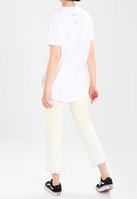 Kurzes weißes T-Shirt mit abgerundetem Saum und Rückendruck, kombiniert mit hellbeigen, knöchellangen Hosen mit fransigen Kanten und schwarzen Sneakers.