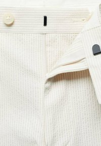 Gros plan de la ceinture d’un pantalon en tissu texturé crème montrant un rabat ouvert avec une fermeture éclair en dessous et un bouton unique au-dessus.