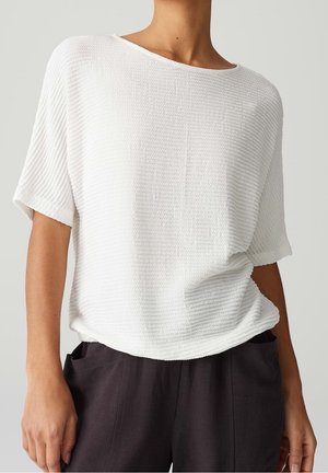 Femme portant un haut blanc texturé à manches courtes associé à un pantalon noir ample avec des poches avant visibles.