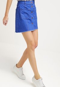 Žena v modré džínové sukni s knoflíky a velkými předními kapsami, v bílé tenisce, prochází před jednobarevným pozadím.