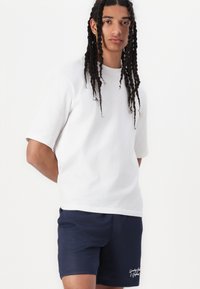 Weißes T-Shirt mit Kurzarm und lockerer Passform, kombiniert mit navyfarbenen, atmungsaktiven Shorts mit subtiler gestickter Details.