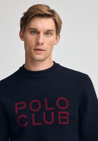 Junger Mann mit hellbraunen Haaren, der einen dunkelblauen Pullover mit rotem "POLO CLUB"-Schriftzug auf der Vorderseite trägt, vor einem einfarbigen Hintergrund.