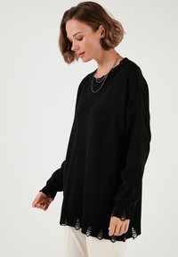 Pulover negru supradimensionat cu mâneci lungi. Are manșete ribbate și un tiv zimțat, decupat. Textură netedă, design simplu.
