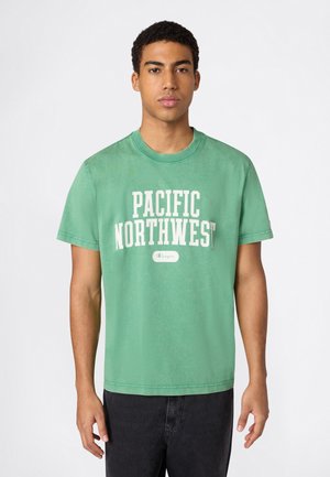 Młody mężczyzna w zielonej koszulce z napisem "PACIFIC NORTHWEST" i czarnych spodniach, stojący na tle jednolitego białego tła.