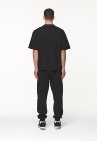Osoba stojí zády k kameře, oblečená v černém oversized tričku a odpovídajících teplácích, na jednobarevném bílém pozadí.
