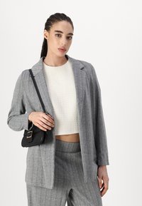 Pilkos spalvos houndstooth švarkas, turintis laisvą siluetą, derinamas su struktūrizuotu baltonu. Modelis laiko mažą juodą rankinę. Matomos pritaikytos kelnės.