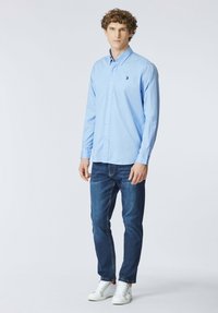 Giovane uomo in piedi, indossa una camicia azzurra con bottoni, jeans blu scuro e sneaker bianche, su uno sfondo chiaro e semplice.