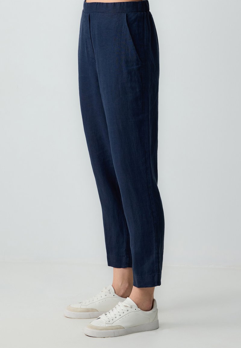 Pantalons en lin bleu marine avec une taille élastique, jambe fuselée et poches latérales, associés à des baskets blanches. Texture lisse.