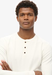 Langärmliges, cremefarbenes Henley-Shirt mit drei braunen Knöpfen am Ausschnitt. Der Stoff wirkt weich mit einer leichten Textur.