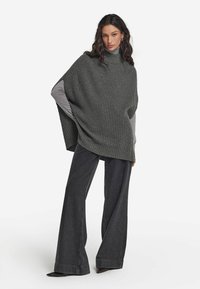 Grå randig poncho med hög krage, lagd över en ljusgrå topp. Pairad med utställda mörk denimjeans och spetsiga svarta klackar.