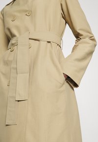 Trench-coat beige à double boutonnage, grands boutons, taille ceinturée et poches latérales. Tissu lisse avec des détails de coutures visibles.