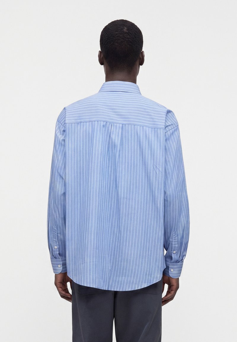 Homme portant une chemise à manches longues rayée bleue et blanche, vu de dos sur un fond clair uni.