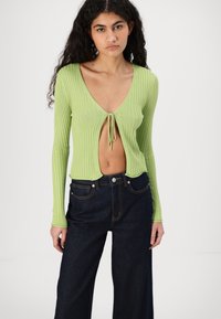 Top acanalado color verde lima con un escote en V profundo y cierre en la parte frontal. Mangas largas y diseño corto combinado con pantalones de mezclilla oscuros de tiro alto.