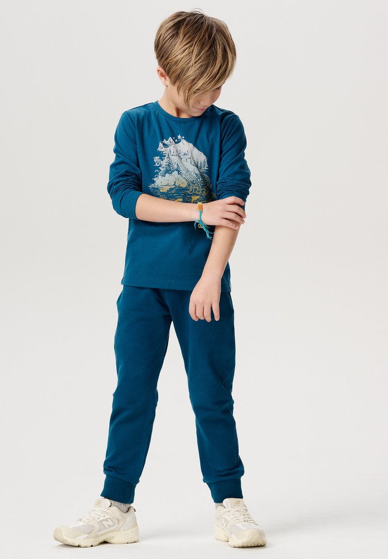 Türaune Langarmshirt mit Berggrafik, kombiniert mit passenden Jogginghosen. Weiße Sneakers mit Mesh-Details runden das Outfit ab.