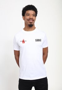 Hvid bomulds t-shirt med "SCARFACE" tekst i sort og et rigtigt flammegrafik i rødt. Kortærmet og rund hals design. Simple og klassisk pasform.
