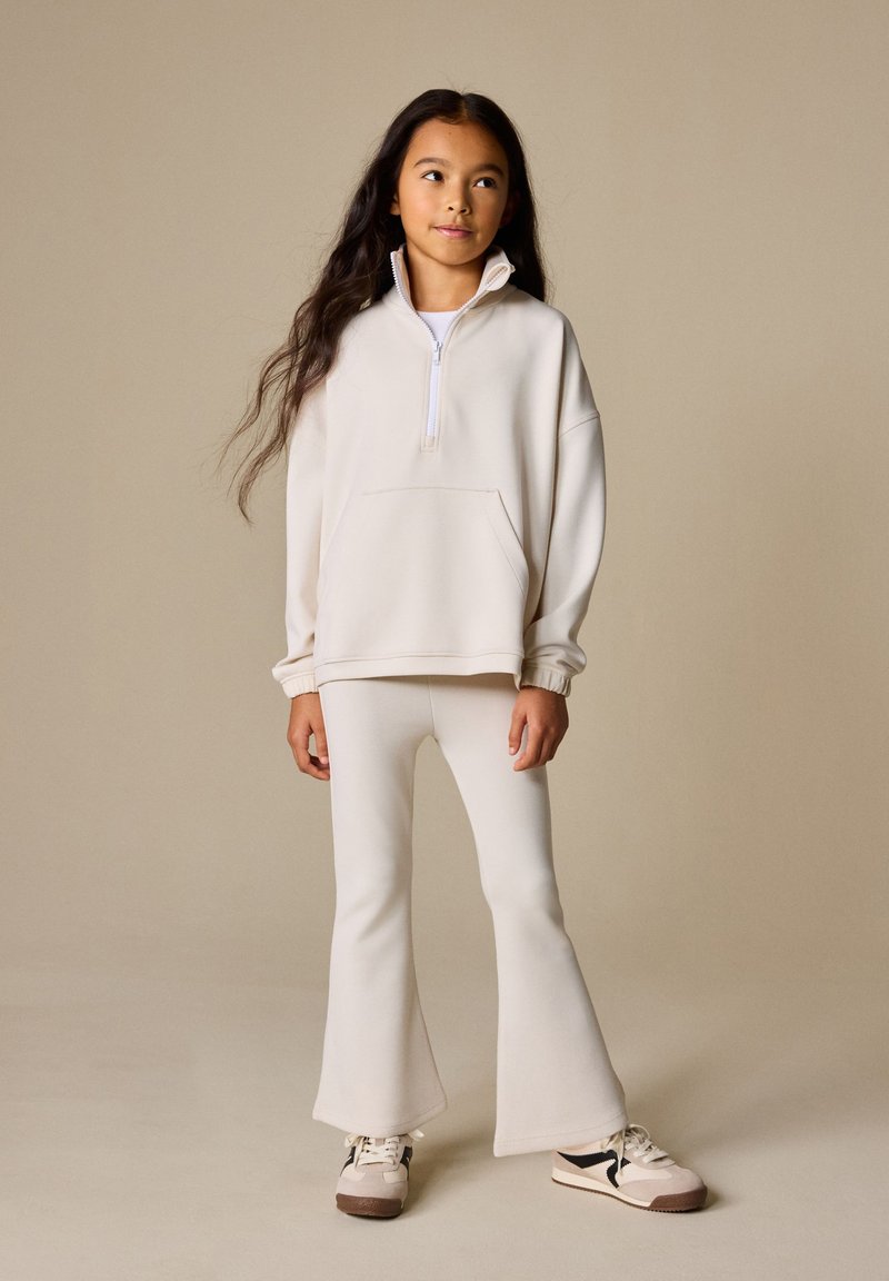 Jeune fille aux longs cheveux portant un sweat crème zippé, un pantalon évasé assorti et des baskets, debout devant un fond beige.