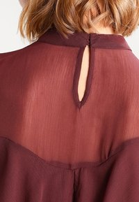 Blouse bordeaux à texture transparente, avec un détail en forme de trou de serrure dans le dos et un col montant. L'ourlet est légèrement arrondi, créant une silhouette fluide.