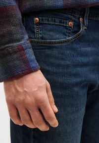 Ανδρικό χέρι που ακουμπά κοντά στην μπροστινή τσέπη σκούρων μπλε τζιν, φοράει πολύχρωμο πουκάμισο με καρό σχέδιο και μακριά μανίκια.