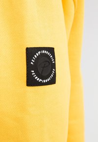 Sudadera amarilla con una textura suave, que presenta un parche rectangular negro con texto blanco y un logo circular.