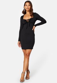 Zwarte lange mouw jurk met een vierkante halslijn en een gestrikt detail aan de voorkant. Gladde textuur en getailleerde pasvorm. Draag met bandjes en versierde hakken.