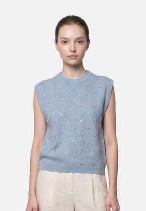 Giovane donna dalla pelle chiara e capelli castani raccolti, che indossa un maglione smanicato azzurro chiaro con motivo a rombi e pantaloni beige chiaro.