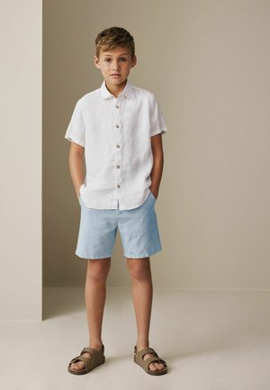 Boy wearing a white linen short-sleeve button-up shirt, light blue shorts, and brown sandals, standing with hands in pockets against a neutral background.