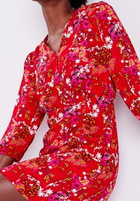 Robe fleurie rouge à manches trois-quarts, avec un décolleté en V, des motifs floraux vibrants en rose et blanc, et des fronces à la taille.
